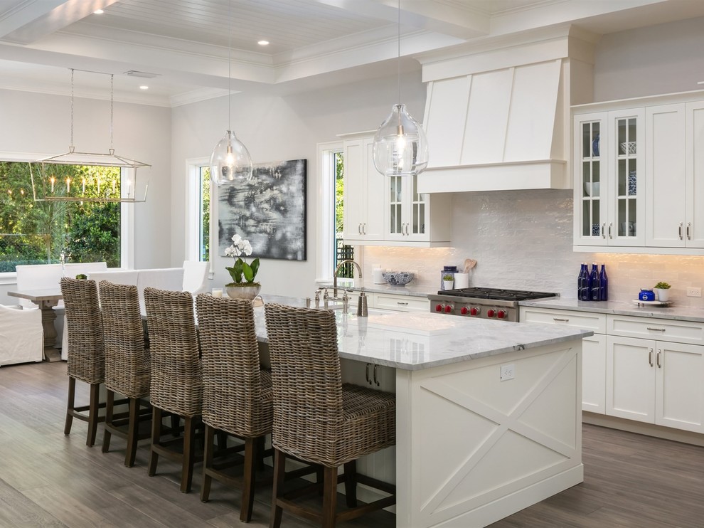 Mid-sized beach style l-shaped open plan kitchen in Orlando with shaker cabinets, white cabinets, stainless steel appliances, with island, white benchtop, white splashback, marble benchtops, dark hardwood floors, a farmhouse sink, stone tile splashback and brown floor.