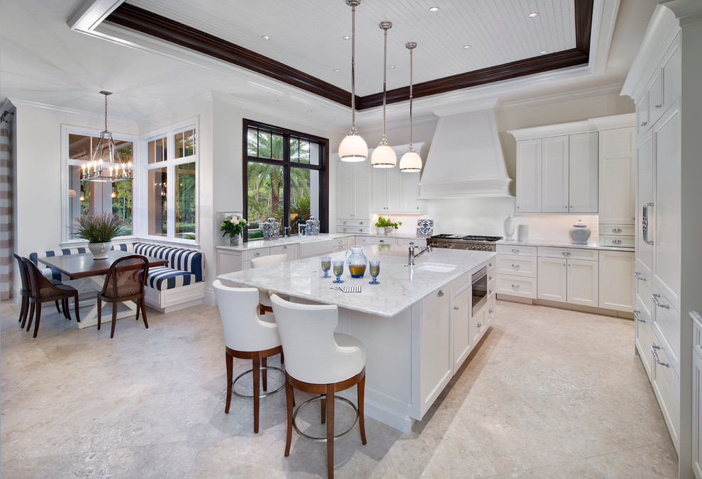 This is an example of a large tropical l-shaped eat-in kitchen in Miami with a farmhouse sink, shaker cabinets, white cabinets, marble benchtops, white splashback, stone slab splashback, panelled appliances, limestone floors, with island and beige floor.
