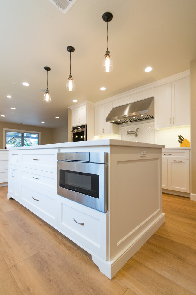 Inspiration for a large traditional u-shaped eat-in kitchen in San Francisco with a farmhouse sink, recessed-panel cabinets, white cabinets, quartz benchtops, white splashback, ceramic splashback, stainless steel appliances, medium hardwood floors, multiple islands and brown floor.