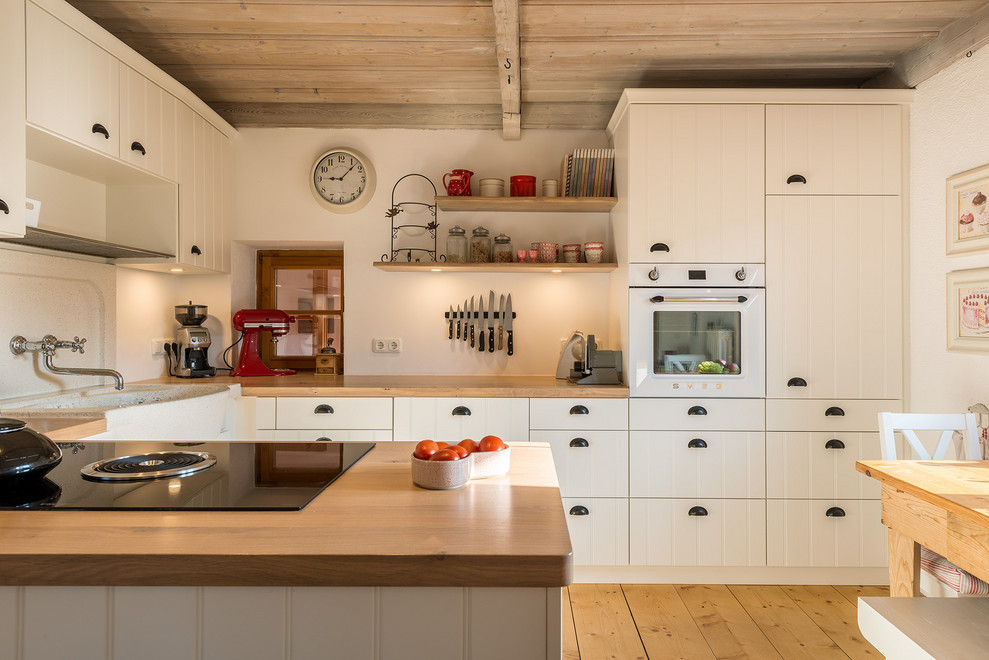 Small country eat-in kitchen in Munich with a farmhouse sink, white cabinets, wood benchtops, white appliances, medium hardwood floors, a peninsula, brown floor, brown benchtop and flat-panel cabinets.