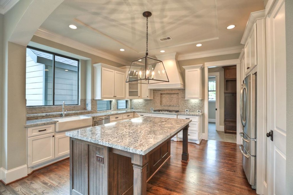 Photo of a large arts and crafts u-shaped separate kitchen in Houston with a farmhouse sink, recessed-panel cabinets, white cabinets, granite benchtops, beige splashback, stone tile splashback, stainless steel appliances, medium hardwood floors and with island.