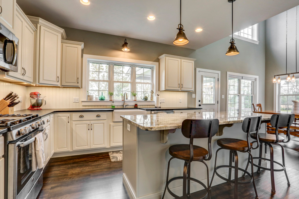 Photo of a country l-shaped eat-in kitchen with a farmhouse sink, white cabinets, granite benchtops, white splashback, shiplap splashback, stainless steel appliances, vinyl floors, with island, brown floor and black benchtop.