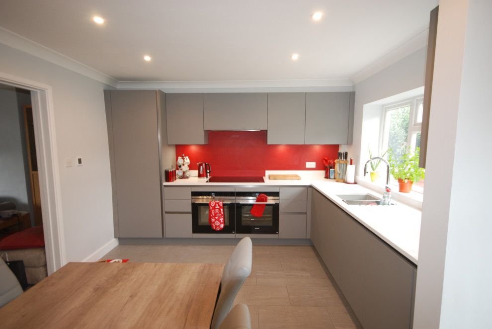 Inspiration for a mid-sized contemporary l-shaped eat-in kitchen in Surrey with an undermount sink, flat-panel cabinets, grey cabinets, solid surface benchtops, red splashback, timber splashback, black appliances, porcelain floors, no island, grey floor and white benchtop.