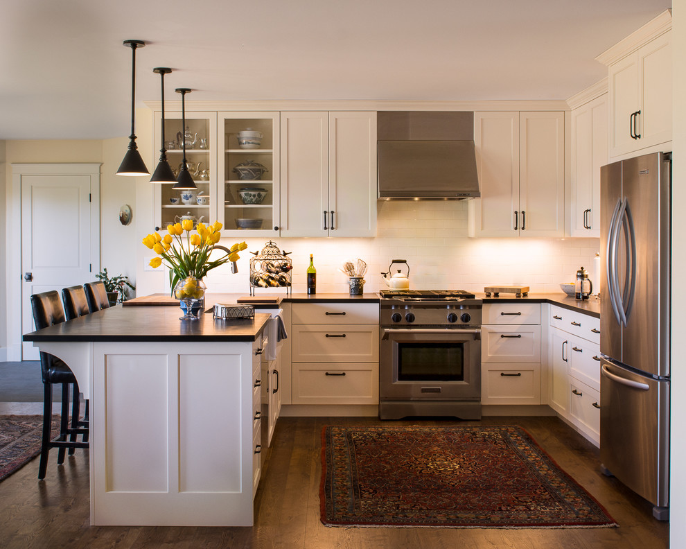 Example of a transitional u-shaped dark wood floor kitchen design with a farmhouse sink, shaker cabinets, white cabinets, white backsplash, subway tile backsplash, stainless steel appliances and a peninsula