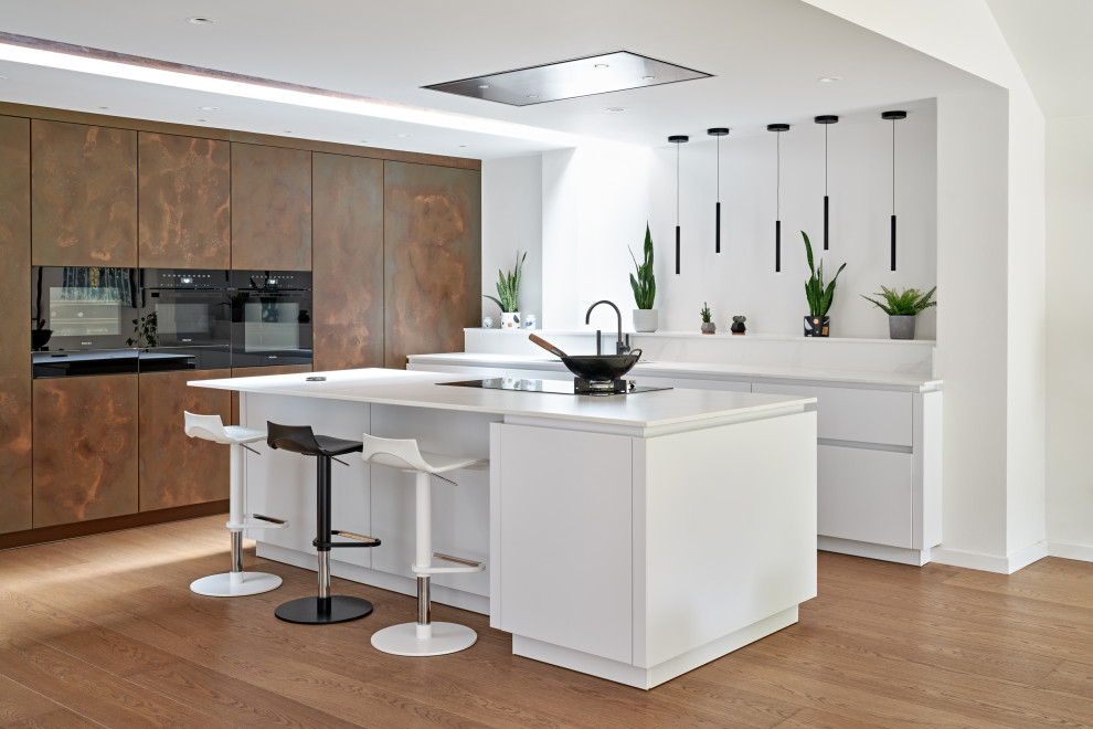 Large contemporary l-shaped open plan kitchen in London with an integrated sink, flat-panel cabinets, composite countertops, black appliances, light hardwood flooring, an island, brown floors, white worktops and feature lighting.