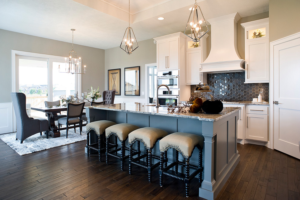 Example of a mid-sized transitional l-shaped medium tone wood floor and brown floor eat-in kitchen design with a farmhouse sink, raised-panel cabinets, white cabinets, granite countertops, gray backsplash, glass tile backsplash, paneled appliances and an island