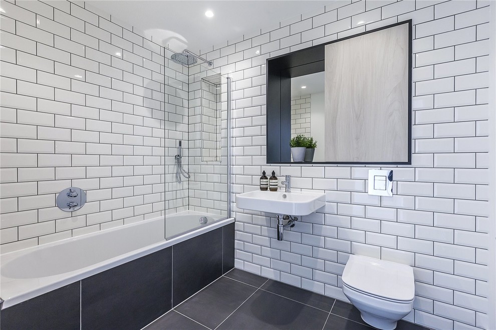 This is an example of a mid-sized modern kids bathroom in London with recessed-panel cabinets, medium wood cabinets, a drop-in tub, a curbless shower, a wall-mount toilet, gray tile, ceramic tile, white walls, ceramic floors, a wall-mount sink, tile benchtops, grey floor, an open shower and white benchtops.