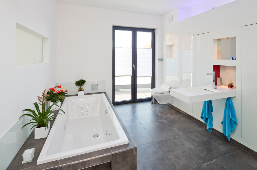 This is an example of a mid-sized modern master bathroom in Hamburg with flat-panel cabinets, white cabinets, a drop-in tub, a curbless shower, gray tile, ceramic tile, white walls, ceramic floors, an integrated sink, solid surface benchtops and a wall-mount toilet.
