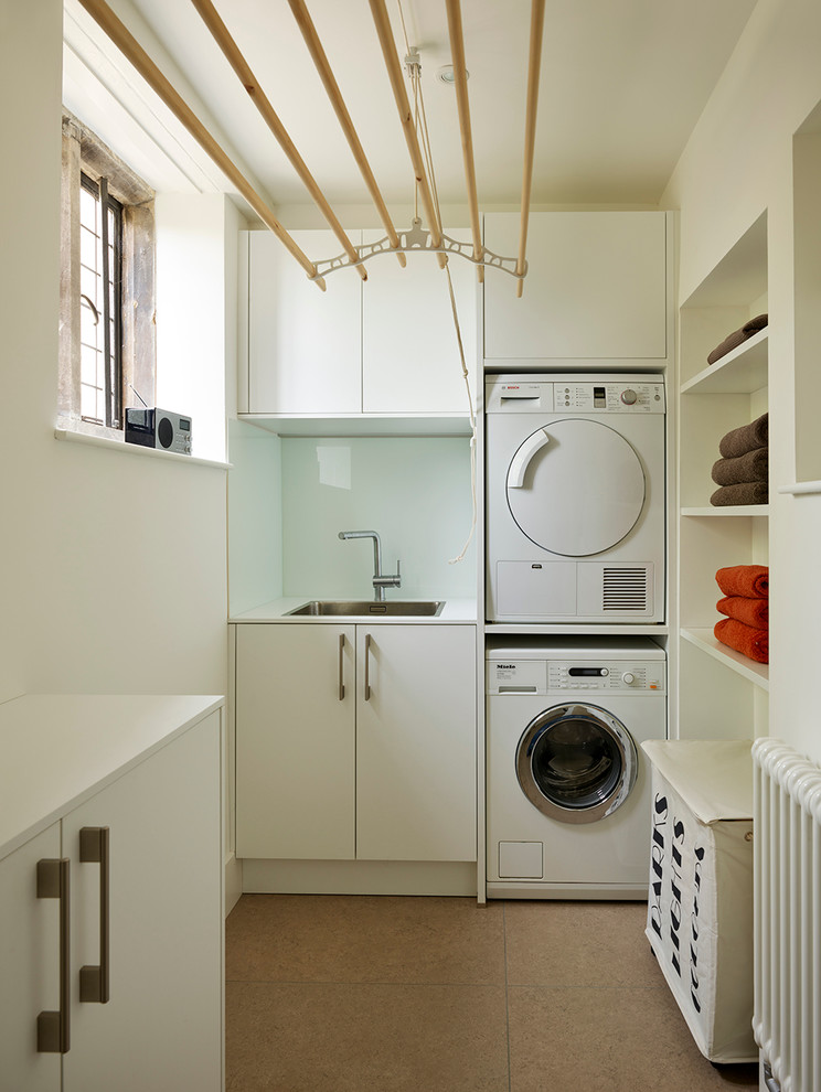 Photo of a mid-sized contemporary u-shaped utility room in Other with an integrated sink, flat-panel cabinets, white cabinets, laminate benchtops, white walls, porcelain floors and a stacked washer and dryer.