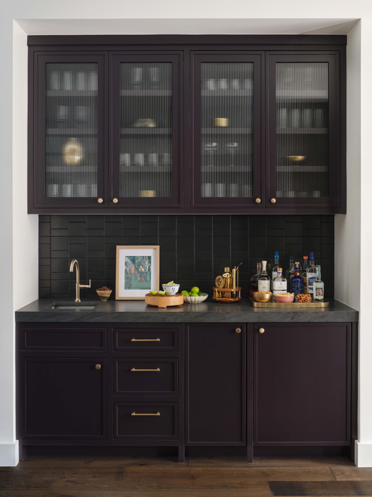 Mid-sized transitional single-wall medium tone wood floor and brown floor wet bar photo in Austin with a drop-in sink, glass-front cabinets, soapstone countertops, black backsplash and black countertops