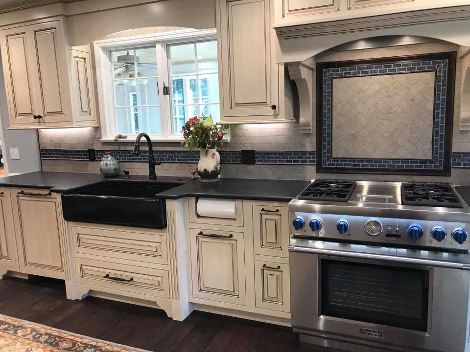 Photo of a mid-sized country l-shaped eat-in kitchen in Other with a farmhouse sink, distressed cabinets, stainless steel appliances, raised-panel cabinets, soapstone benchtops, grey splashback, ceramic splashback, dark hardwood floors, with island and brown floor.