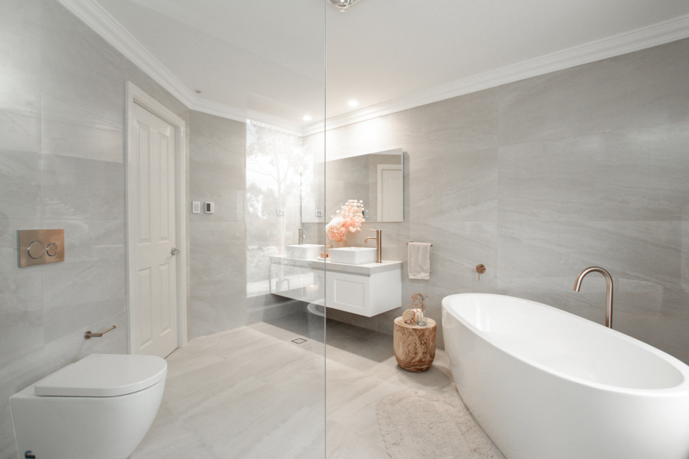 Inredning av ett modernt stort en-suite badrum, med skåp i shakerstil, vita skåp, ett fristående badkar, en öppen dusch, en toalettstol med hel cisternkåpa, grå kakel, grå väggar, ett fristående handfat, grått golv och med dusch som är öppen