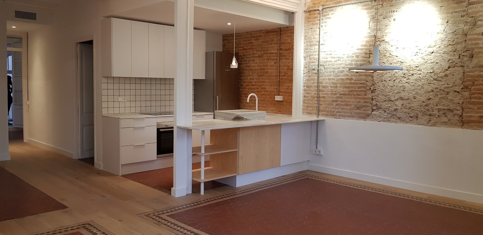 Photo of a mid-sized industrial u-shaped eat-in kitchen in Barcelona with a farmhouse sink, flat-panel cabinets, white cabinets, marble benchtops, white splashback, subway tile splashback, stainless steel appliances, terra-cotta floors, a peninsula, red floor and white benchtop.
