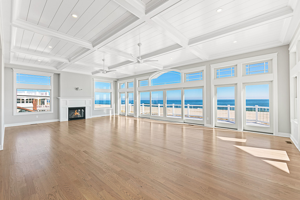Expansive beach style open concept living room in Other with grey walls, light hardwood floors, a standard fireplace, a wood fireplace surround, a wall-mounted tv, beige floor and coffered.