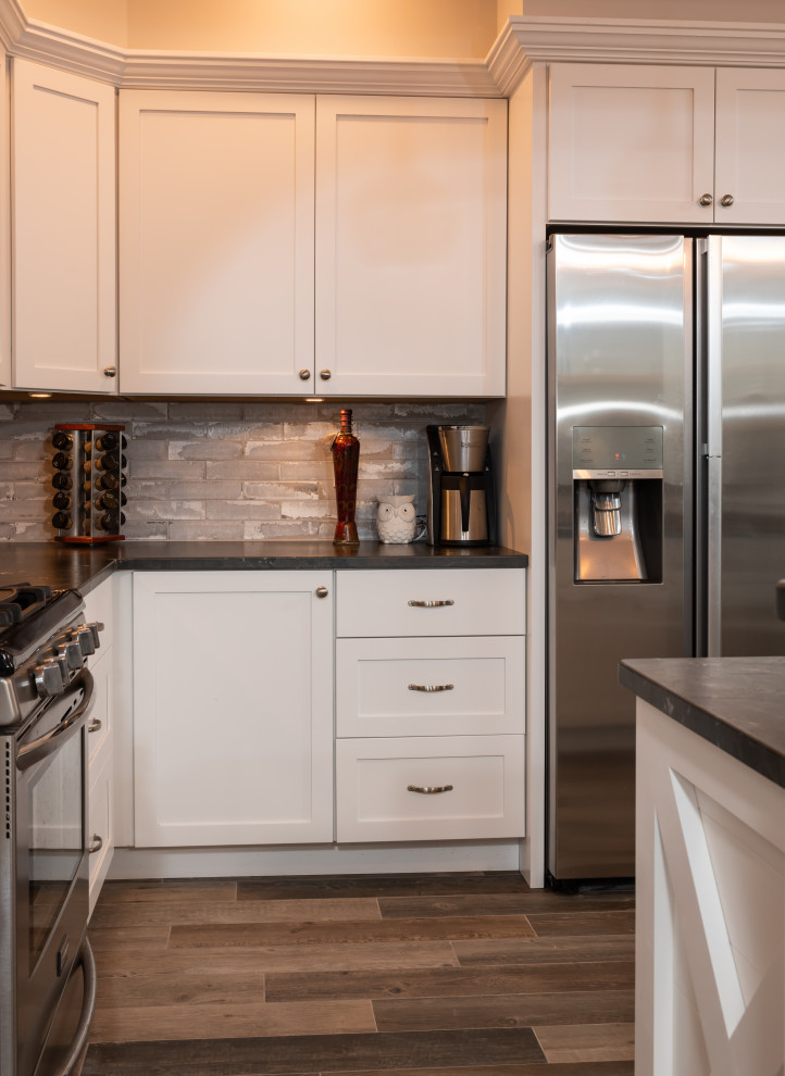 Example of a mid-sized mountain style l-shaped porcelain tile and gray floor open concept kitchen design with an undermount sink, shaker cabinets, white cabinets, granite countertops, gray backsplash, brick backsplash, stainless steel appliances, an island and black countertops