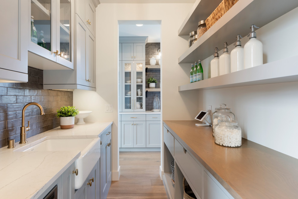 This is an example of a transitional galley kitchen pantry in Minneapolis with a farmhouse sink, shaker cabinets, grey cabinets, brown splashback, subway tile splashback, light hardwood floors, beige floor and white benchtop.