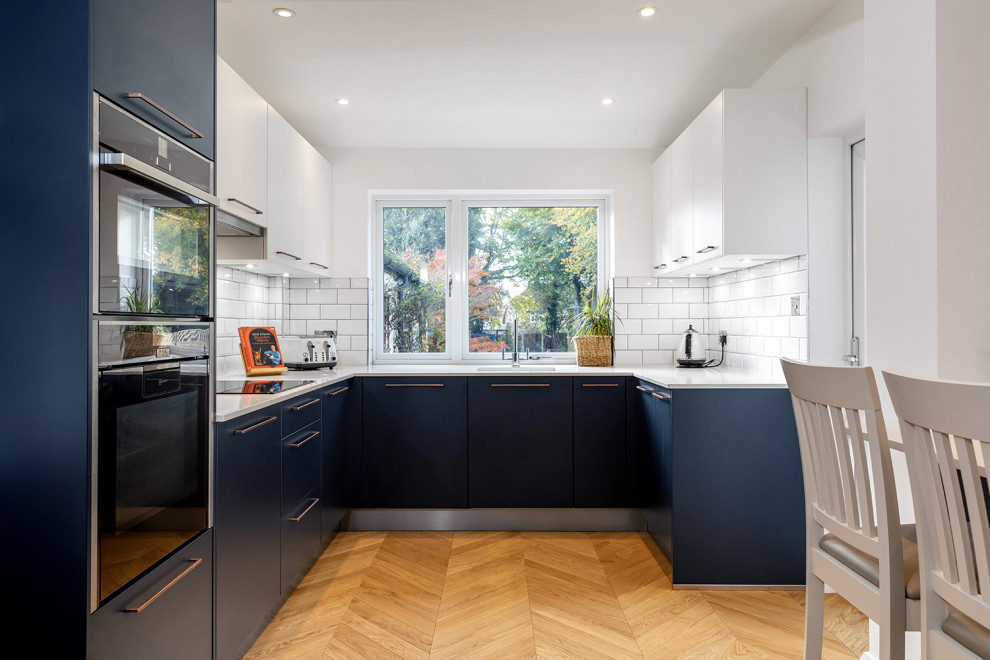 Photo of a mid-sized contemporary u-shaped eat-in kitchen in Hertfordshire with an undermount sink, flat-panel cabinets, blue cabinets, quartzite benchtops, white splashback, ceramic splashback, black appliances, light hardwood floors, with island, brown floor and white benchtop.