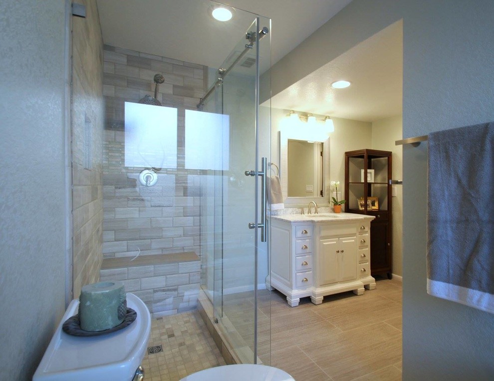 This is an example of a large transitional master bathroom in Denver with raised-panel cabinets, white cabinets, a drop-in tub, a corner shower, a one-piece toilet, beige tile, stone tile, beige walls, porcelain floors, marble benchtops and an undermount sink.