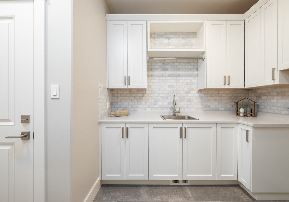 Photo of a large traditional l-shaped dedicated laundry room in Other with a drop-in sink, shaker cabinets, white cabinets, solid surface benchtops, grey walls, concrete floors, a side-by-side washer and dryer, grey floor and white benchtop.