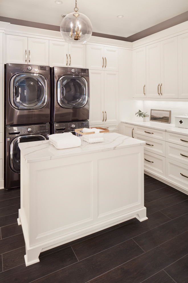 Large transitional l-shaped dedicated laundry room in Milwaukee with recessed-panel cabinets, white cabinets, white splashback, grey walls, a stacked washer and dryer, brown floor and white benchtop.