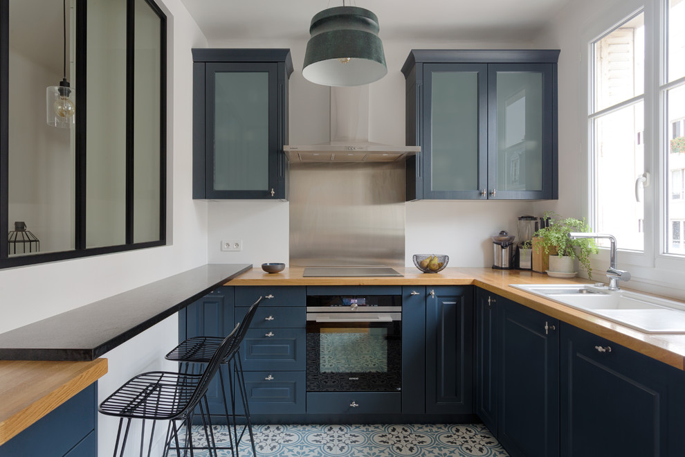 This is an example of a contemporary l-shaped separate kitchen in Paris with a triple-bowl sink, raised-panel cabinets, blue cabinets, wood benchtops, metallic splashback, slate splashback, black appliances, multi-coloured floor and brown benchtop.