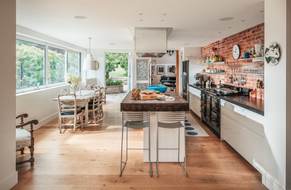 Photo of a large contemporary single-wall eat-in kitchen in Sussex with a drop-in sink, flat-panel cabinets, white cabinets, granite benchtops, brick splashback, black appliances, medium hardwood floors, a peninsula and black benchtop.