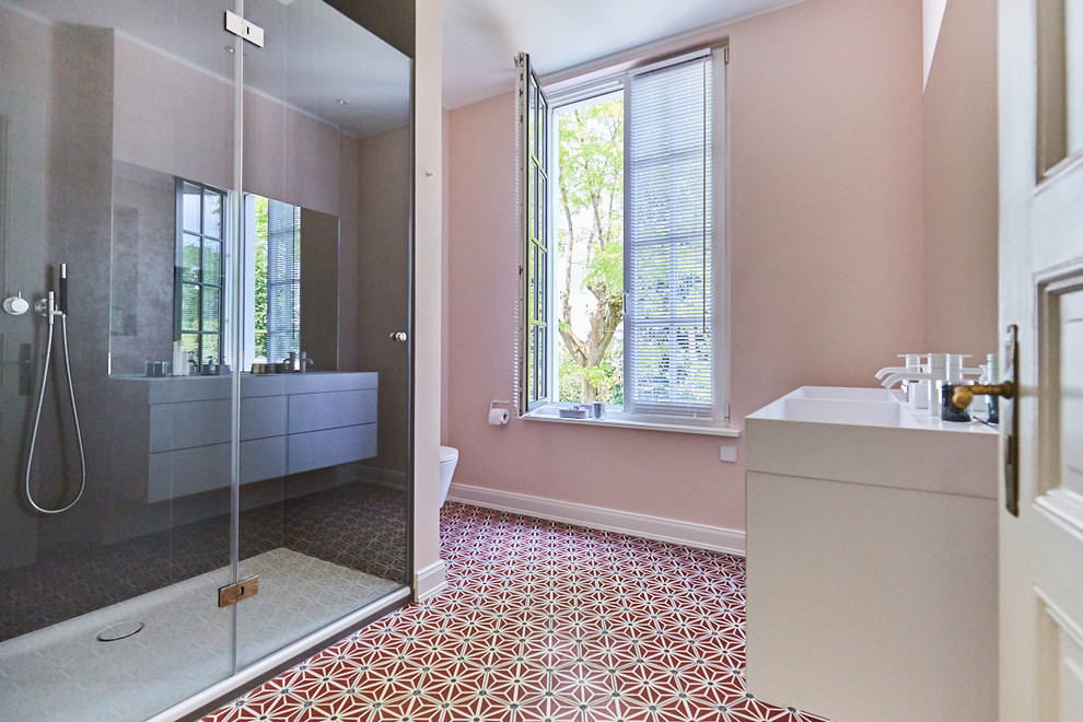 Photo of a mid-sized traditional bathroom in Hamburg with flat-panel cabinets, white cabinets, an alcove shower, a wall-mount toilet, pink walls, cement tiles, an integrated sink, solid surface benchtops, red floor, a hinged shower door and white benchtops.