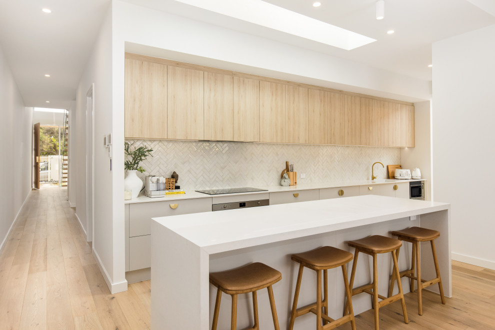 This is an example of an arts and crafts galley kitchen with an undermount sink, flat-panel cabinets, grey cabinets, white splashback, stainless steel appliances, medium hardwood floors, with island, brown floor and white benchtop.