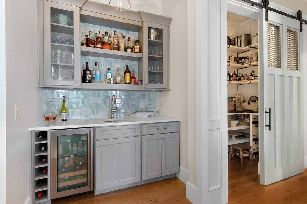 Inspiration for a large beach style galley home bar in Charleston with grey cabinets, quartzite benchtops, blue splashback, mosaic tile splashback, medium hardwood floors, brown floor, white benchtop and a drop-in sink.