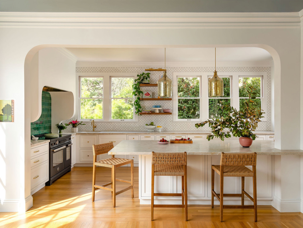 Eat-in kitchen - large transitional l-shaped medium tone wood floor and brown floor eat-in kitchen idea in San Francisco with a farmhouse sink, shaker cabinets, white cabinets, quartzite countertops, white backsplash, marble backsplash, black appliances, a peninsula and beige countertops