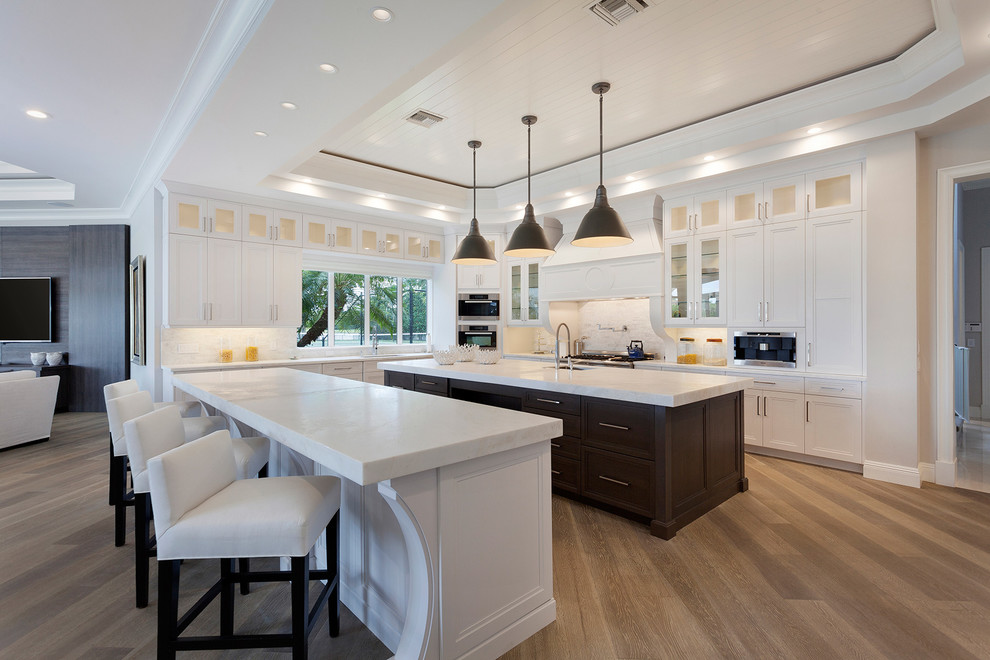 Photo of a transitional u-shaped kitchen in Miami with recessed-panel cabinets, white cabinets, white splashback, stainless steel appliances, medium hardwood floors and multiple islands.