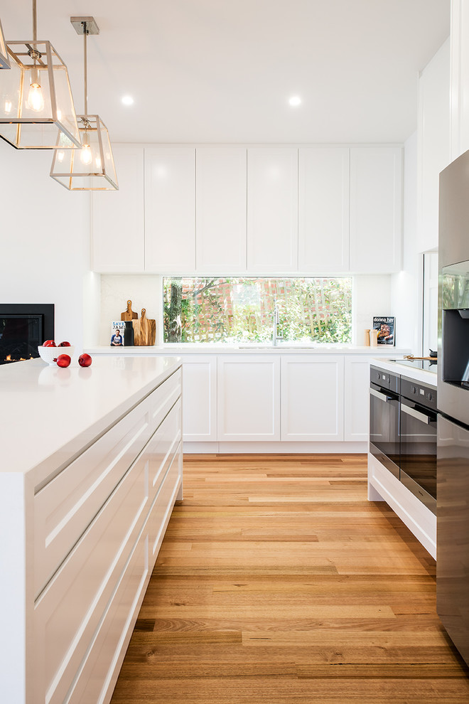 Large contemporary l-shaped open plan kitchen in Melbourne with an undermount sink, shaker cabinets, white cabinets, quartz benchtops, white splashback, window splashback, stainless steel appliances, medium hardwood floors, with island and brown floor.