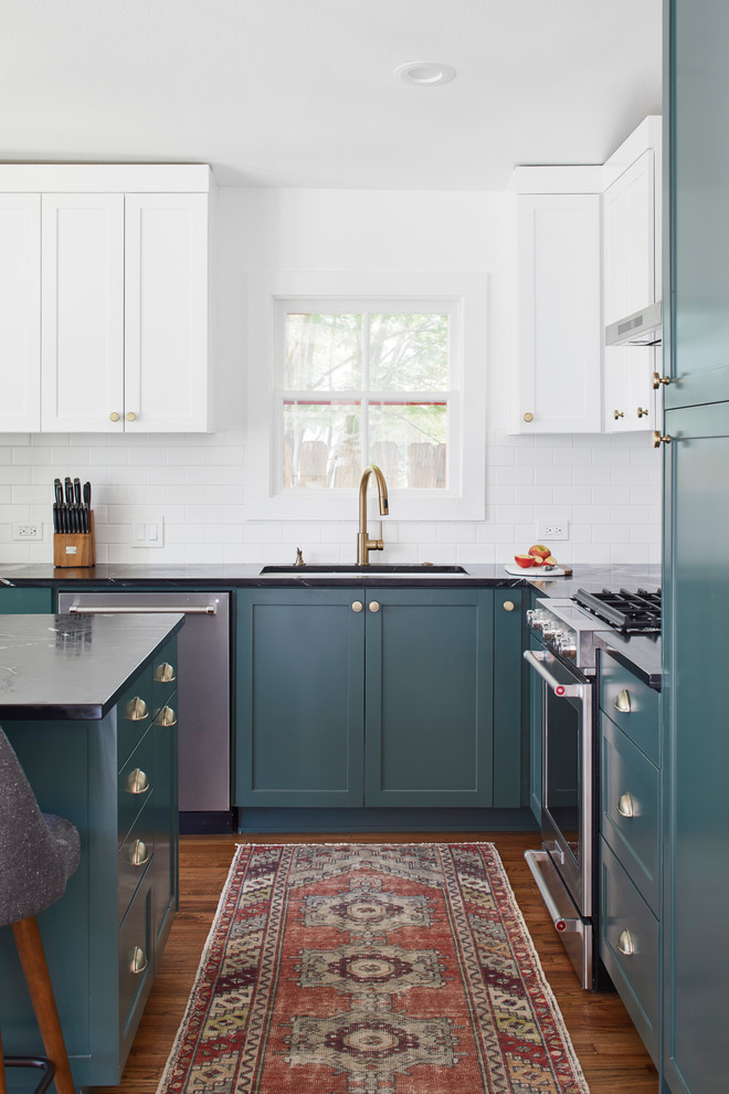 Design ideas for a mid-sized eclectic l-shaped open plan kitchen in Austin with an undermount sink, shaker cabinets, green cabinets, soapstone benchtops, white splashback, ceramic splashback, stainless steel appliances, medium hardwood floors, with island, brown floor and black benchtop.