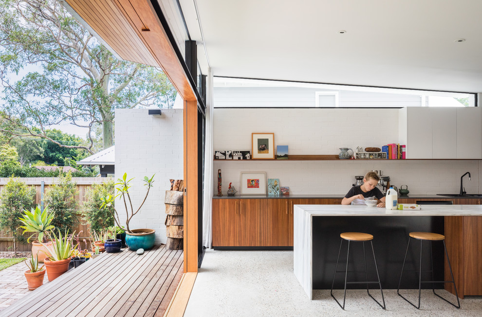Inspiration for a large contemporary galley open plan kitchen in Sydney with a double-bowl sink, flat-panel cabinets, medium wood cabinets, marble benchtops, white splashback, brick splashback, black appliances, concrete floors, with island, grey floor and black benchtop.