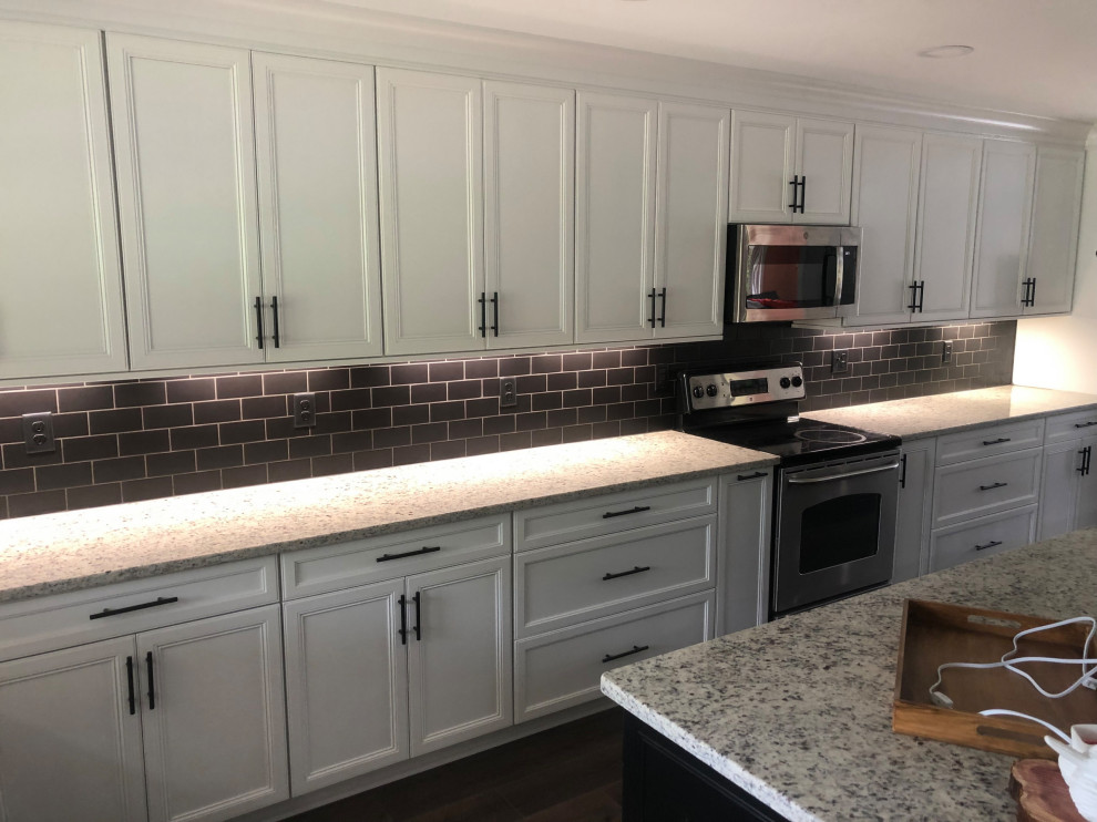 Large modern l-shaped kitchen pantry in Charlotte with a farmhouse sink, shaker cabinets, white cabinets, granite benchtops, brown splashback, subway tile splashback, stainless steel appliances, dark hardwood floors, with island and brown floor.