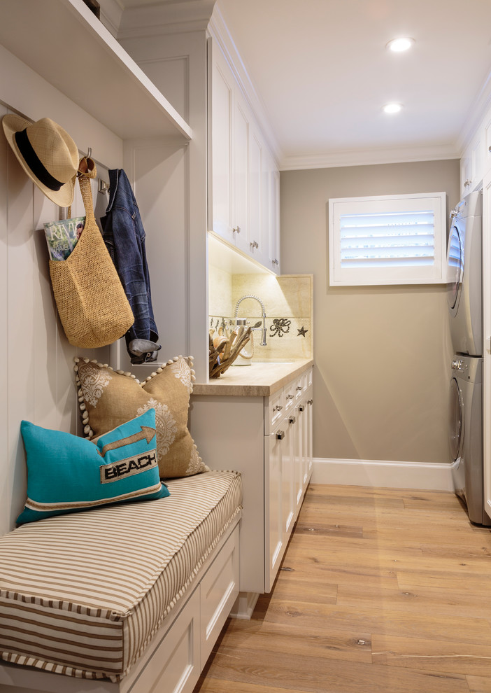 Inspiration for a large beach style galley dedicated laundry room in Orange County with a single-bowl sink, recessed-panel cabinets, white cabinets, limestone benchtops, grey walls, light hardwood floors and a stacked washer and dryer.