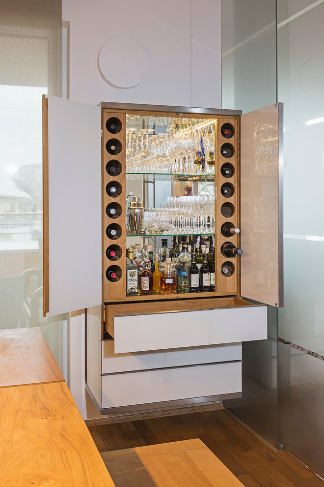 Photo of a small contemporary home bar in Munich with flat-panel cabinets, white cabinets, medium hardwood floors, wood benchtops and mirror splashback.