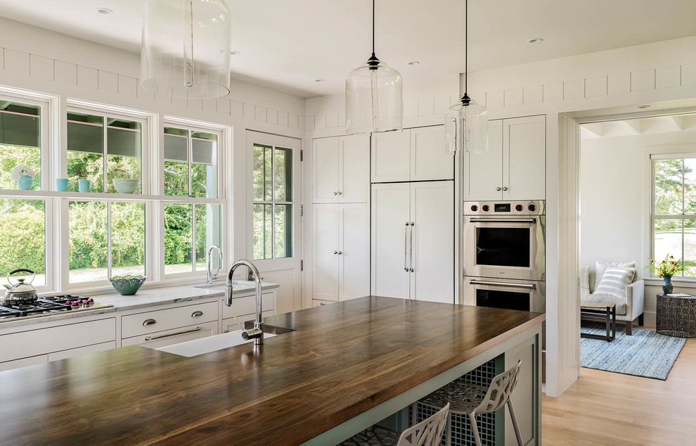 This is an example of a mid-sized beach style galley eat-in kitchen in Portland Maine with a single-bowl sink, shaker cabinets, white cabinets, wood benchtops, white splashback, stainless steel appliances, light hardwood floors and with island.