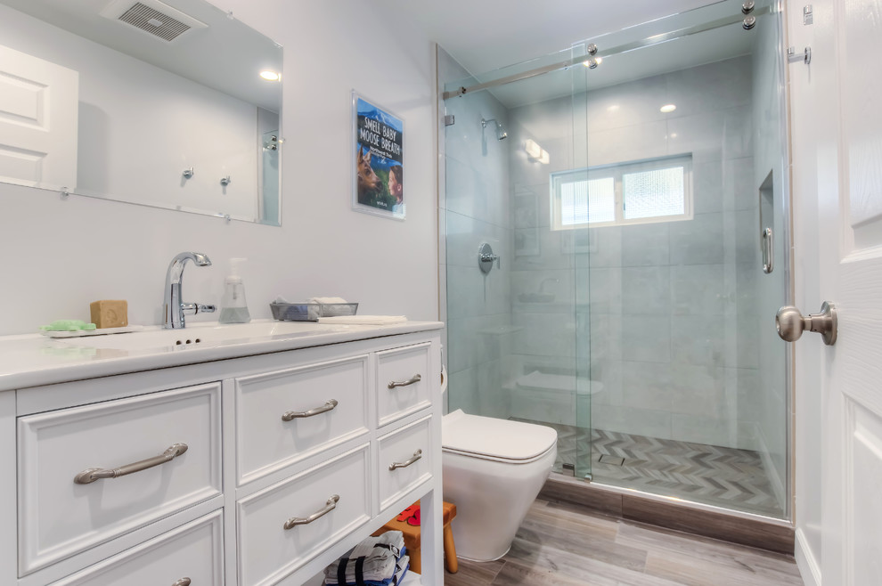 Mid-sized transitional 3/4 bathroom in Los Angeles with white cabinets, white walls, brown floor, a sliding shower screen, recessed-panel cabinets, an alcove shower, a one-piece toilet, gray tile, cement tile, an undermount sink, marble benchtops and laminate floors.