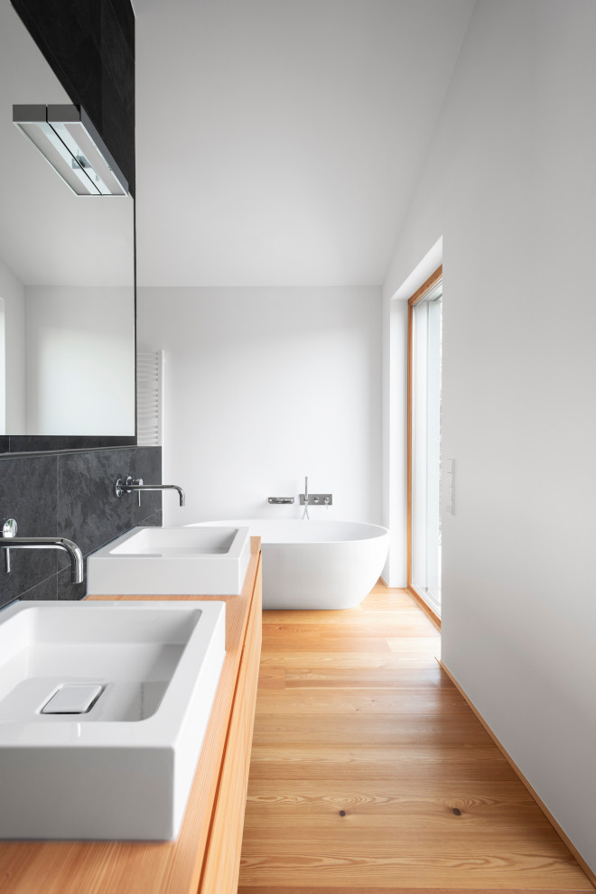 Badezimmer En Suite With flächenbündigen Schrankfronten, hellen Holzschränken, freistehender Badewanne, bodengleicher Dusche, Wandtoilette, schwarzen Fliesen, Schieferfliesen, weißer Wandfarbe, Schieferboden, Aufsatzwaschbecken, Waschtisch aus Holz, schwarzem Boden, Falttür-Duschabtrennung and brauner Waschtischplatte in Munich