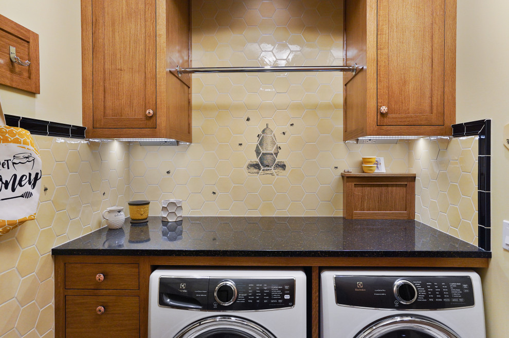 Bild på en stor amerikansk svarta svart tvättstuga, med en allbänk, skåp i shakerstil, skåp i mellenmörkt trä, granitbänkskiva, gult stänkskydd, stänkskydd i keramik, gröna väggar, klinkergolv i porslin och en tvättmaskin och torktumlare bredvid varandra