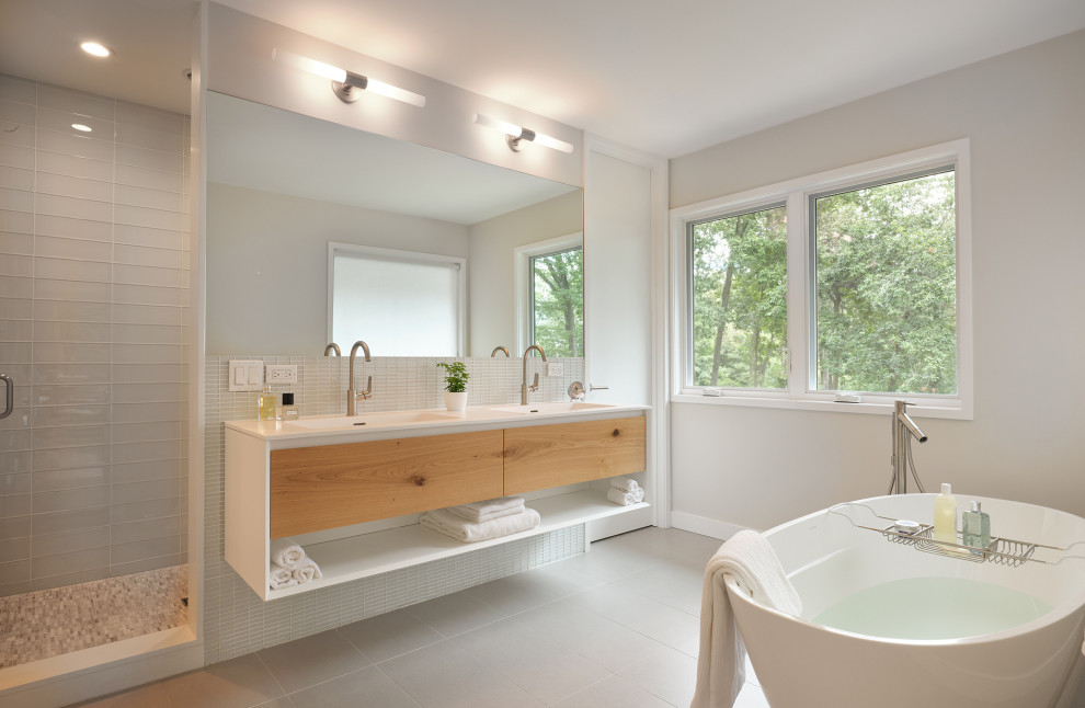 Photo of a mid-sized contemporary master bathroom in New York with flat-panel cabinets, a freestanding tub, white tile, glass tile, porcelain floors, an integrated sink, solid surface benchtops, grey floor, a hinged shower door, white benchtops, medium wood cabinets, an alcove shower, grey walls, a double vanity and a floating vanity.