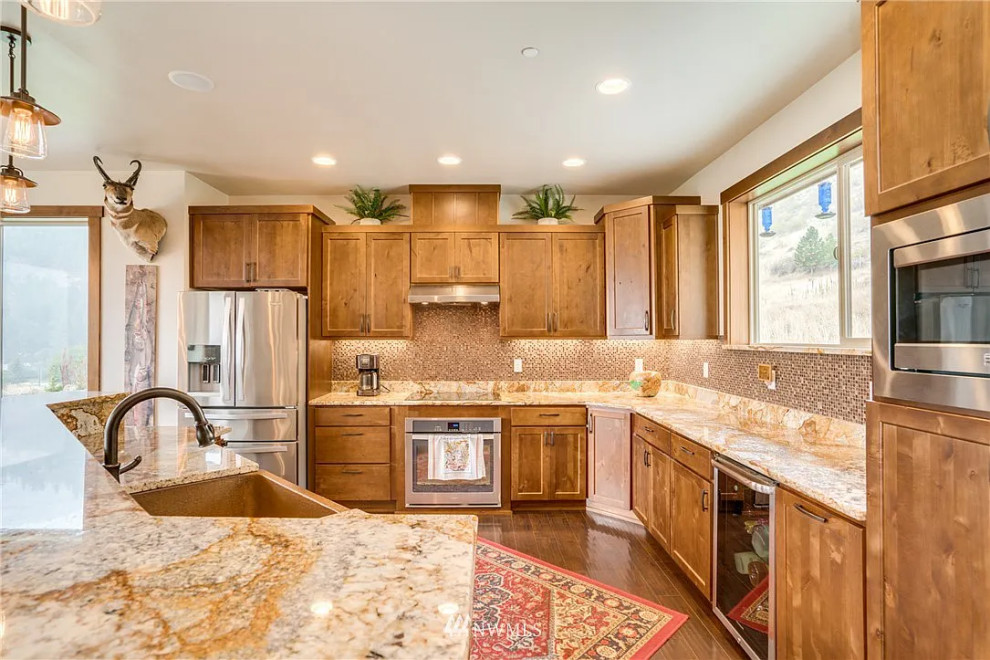 Example of a mid-sized mountain style l-shaped laminate floor, brown floor and tray ceiling open concept kitchen design with a single-bowl sink, shaker cabinets, medium tone wood cabinets, granite countertops, multicolored backsplash, glass tile backsplash, stainless steel appliances, an island and multicolored countertops