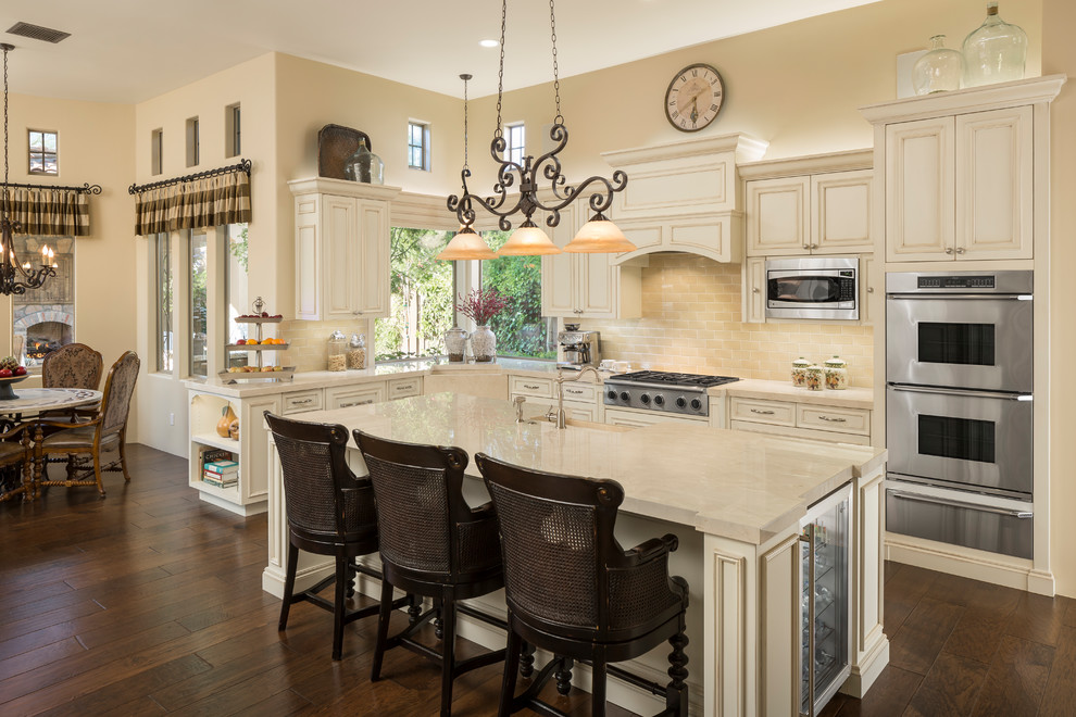 Design ideas for a mid-sized traditional l-shaped eat-in kitchen in Phoenix with recessed-panel cabinets, beige cabinets, beige splashback, subway tile splashback, stainless steel appliances, dark hardwood floors, with island, brown floor, beige benchtop, a farmhouse sink and quartz benchtops.