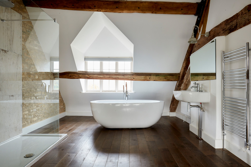 This is an example of a country bathroom in Wiltshire with a freestanding tub, white walls, dark hardwood floors, a wall-mount sink and brown floor.