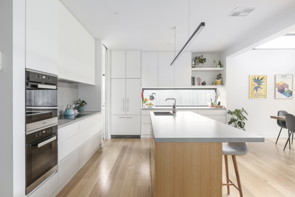 Photo of a contemporary l-shaped kitchen in Melbourne with an undermount sink, flat-panel cabinets, white cabinets, grey splashback, black appliances, light hardwood floors, with island, beige floor and white benchtop.