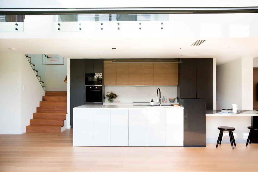 Design ideas for a contemporary galley kitchen in Sunshine Coast with an undermount sink, flat-panel cabinets, light wood cabinets, white splashback, black appliances, light hardwood floors, with island, beige floor and white benchtop.