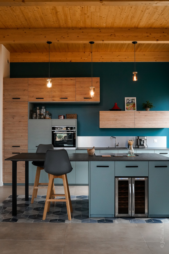 Mid-sized modern galley open plan kitchen in Lyon with an integrated sink, blue cabinets, metallic splashback, stainless steel appliances, ceramic floors, with island, blue floor, grey benchtop and exposed beam.