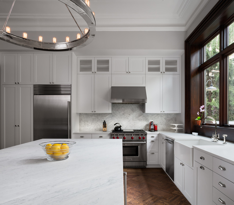Transitional l-shaped eat-in kitchen in New York with a farmhouse sink, shaker cabinets, white cabinets, white splashback, stainless steel appliances, dark hardwood floors, with island, mosaic tile splashback and marble benchtops.