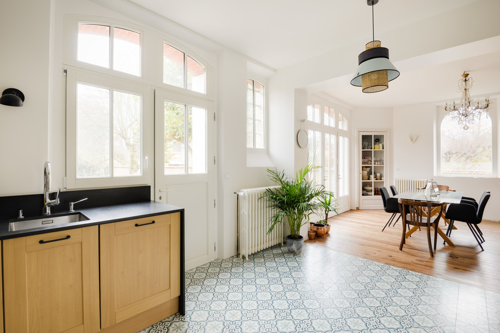 Transitional l-shaped open plan kitchen in Paris with an undermount sink, shaker cabinets, light wood cabinets, stainless steel appliances, cement tiles, blue floor, black benchtop, granite benchtops and mirror splashback.